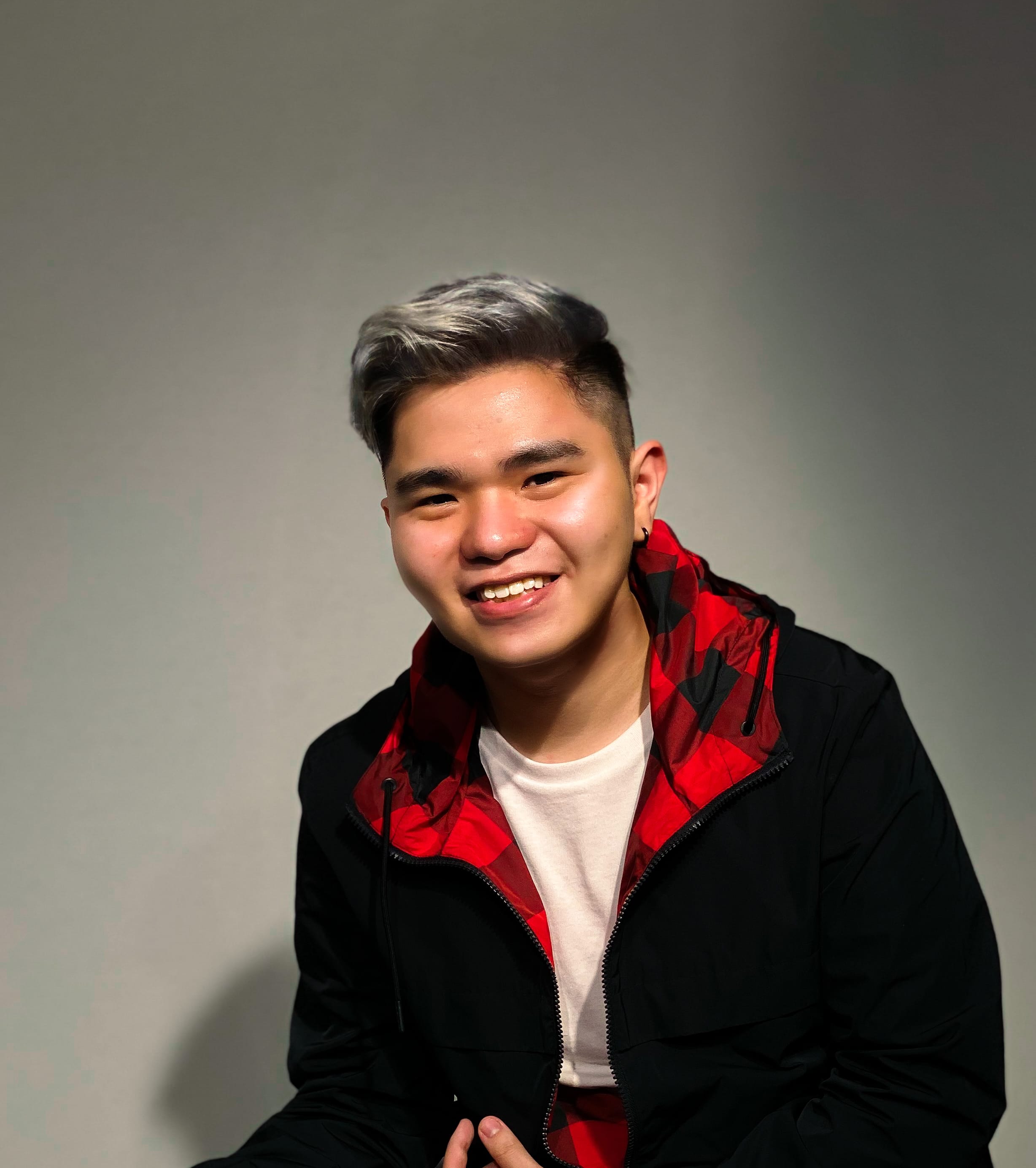 Young man with silver-dyed hair smiling, wearing a black jacket with red plaid hood, against a gray background.